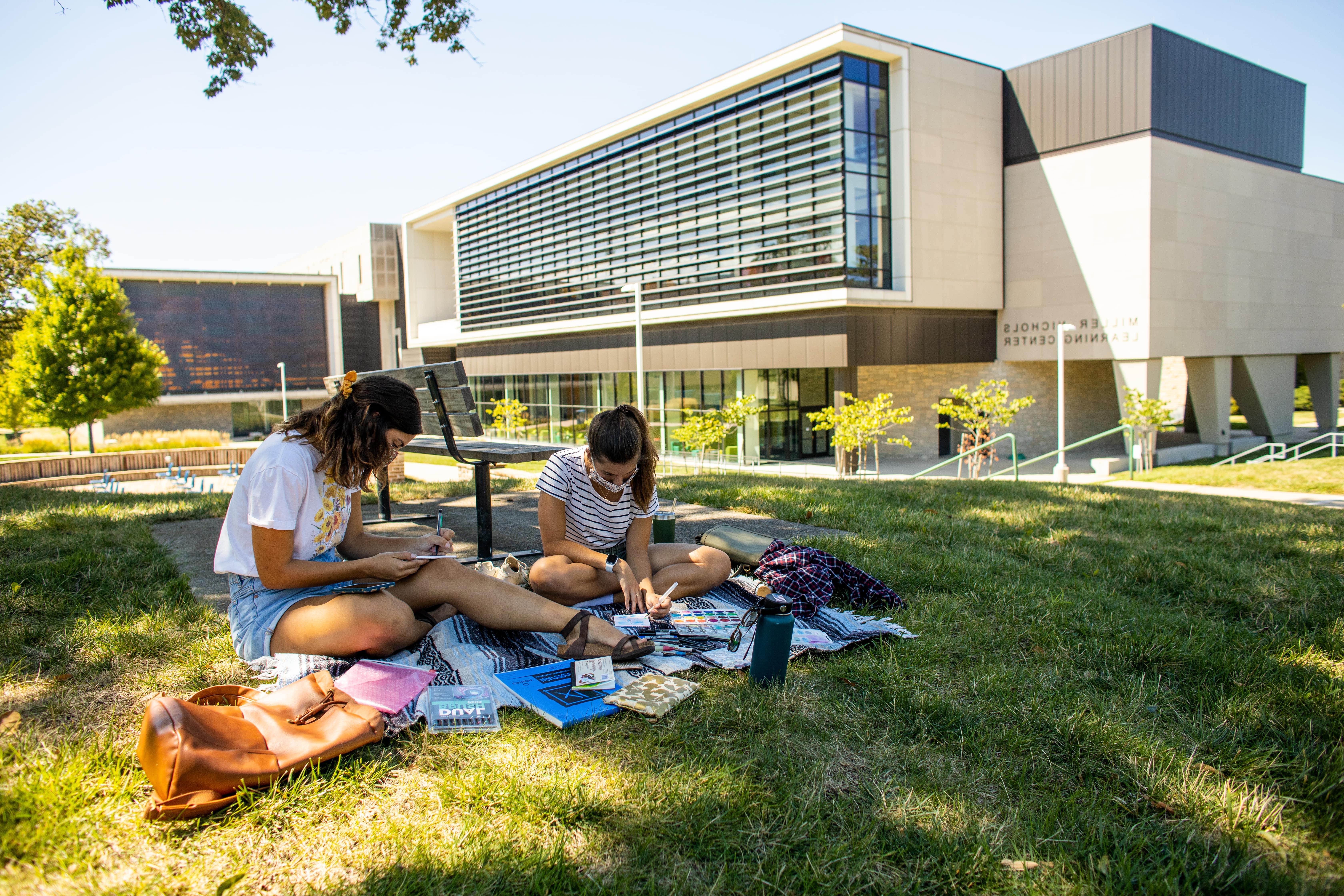 students by the library, painting on a blanket outside on a sunny day.