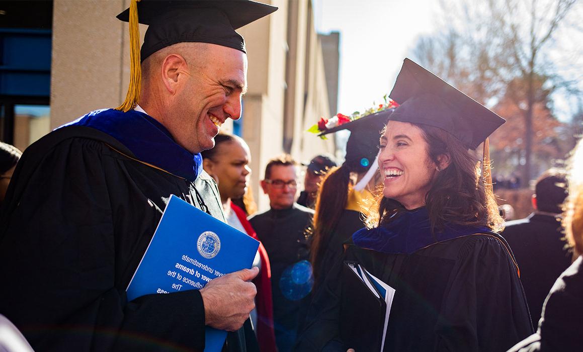 Person presenting as white woman in graduation cap and gown smiles at person presenting as white man wearing cap and gown