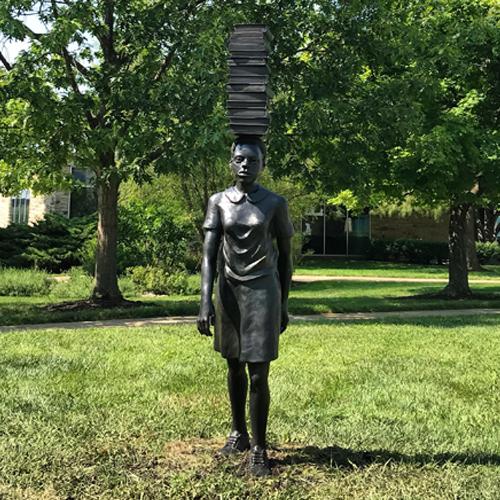 The sculpture by Flávio Cerqueira, titled “Any Word Except Wait” features a female with a stack of books on her head taking a step forward 