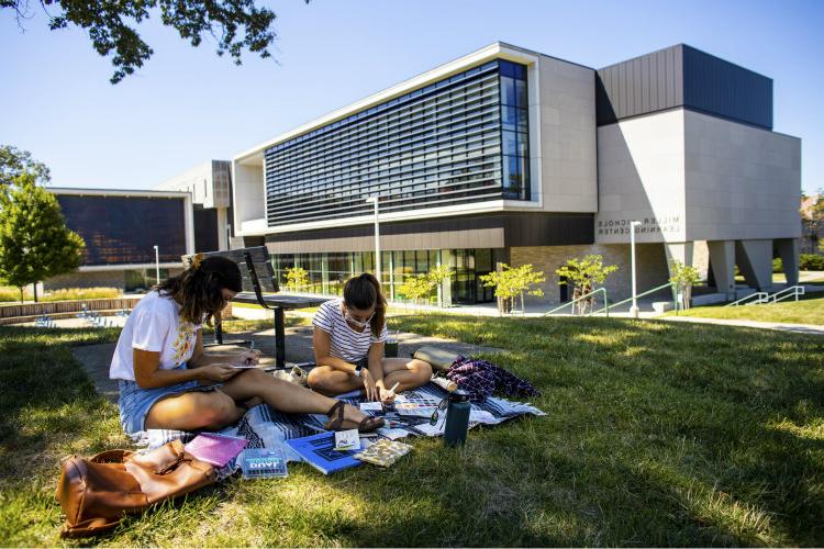 Two masked students on lawn outside library and learning center.