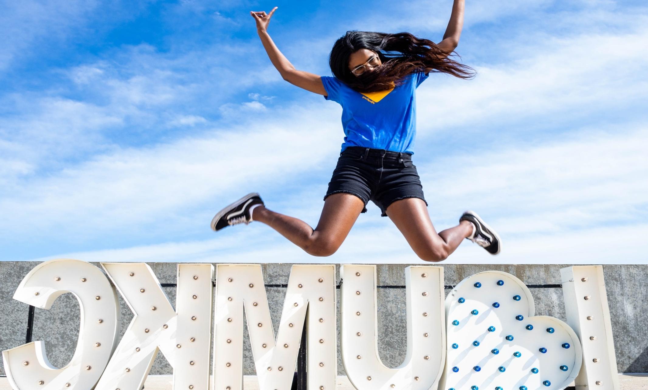 A woman in a blue shirt is jumping in front of a large light-up sign that reads 'I <3 UMKC'