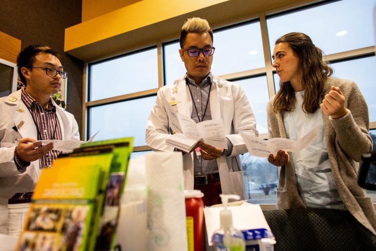 A female talks to two males; health brochures and supplies are in the foreground.