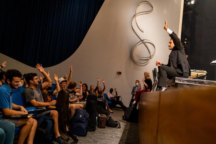 A conservatory professor sits on a stage with his feet dangling addressing students sitting in an auditorium. Nearly all of them have their hand raised. 