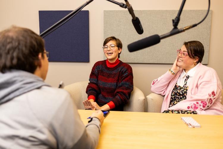 Three students sit around a table smiling with microphones sitting in front of them