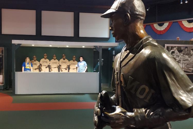 Hunter and Mariah stand in a dugout with a statue of a pitcher in the foreground