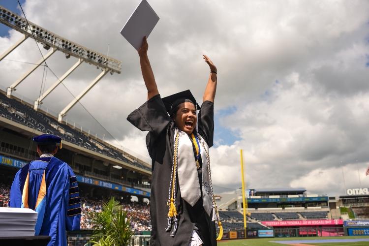 A graduate stands on the stage with her hands in the air, diploma in one hand, smiling