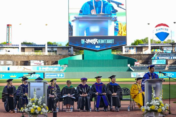 Chancellor Agrawal stands at the podium with the deans seated behind him on the field of Kauffman Stadium