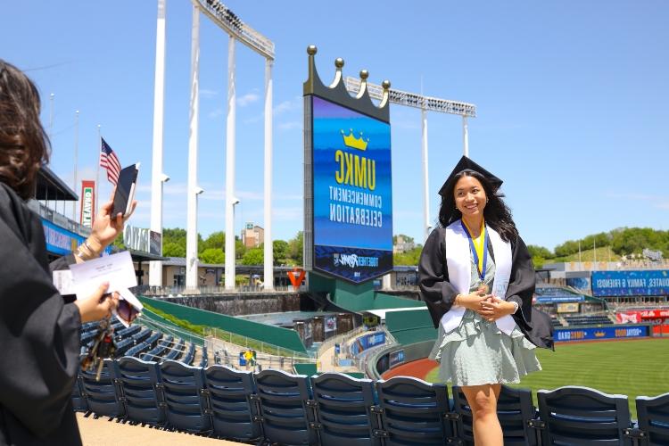 A graduate stands in front of Crown Vision at Kauffman Stadium as someone takes her photograph