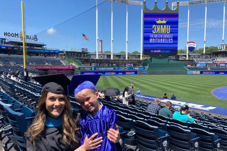 A mother and son in graduation robes pose for a photo in front of Crown Vision at Kauffman Stadium