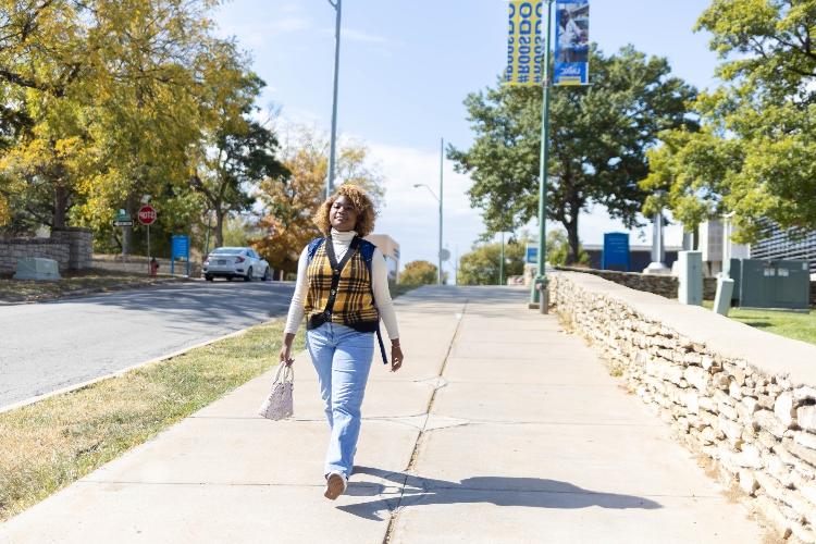 Jayla Williams带着背包和钱包走在UMKC校园的人行道上. In the background you can see UMKC signs.