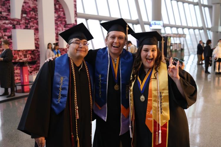 Three graduates smile and one holds a Roo Up inside the rotunda of the T-Mobile Center