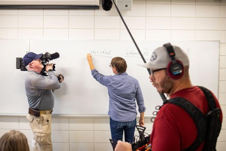 Clancy Martin writes on the whiteboard while teaching a class; CBS cameras and reporters are around him