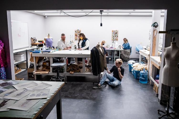 Students and a professor work in a studio. One sews, two look at illustrations on a table and another looks at costumes.