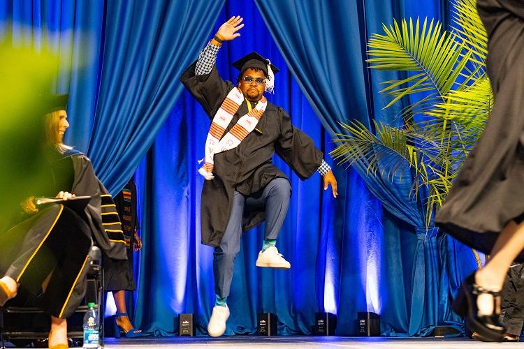 A student is mid-jump as he dances on stage while receiving his diploma