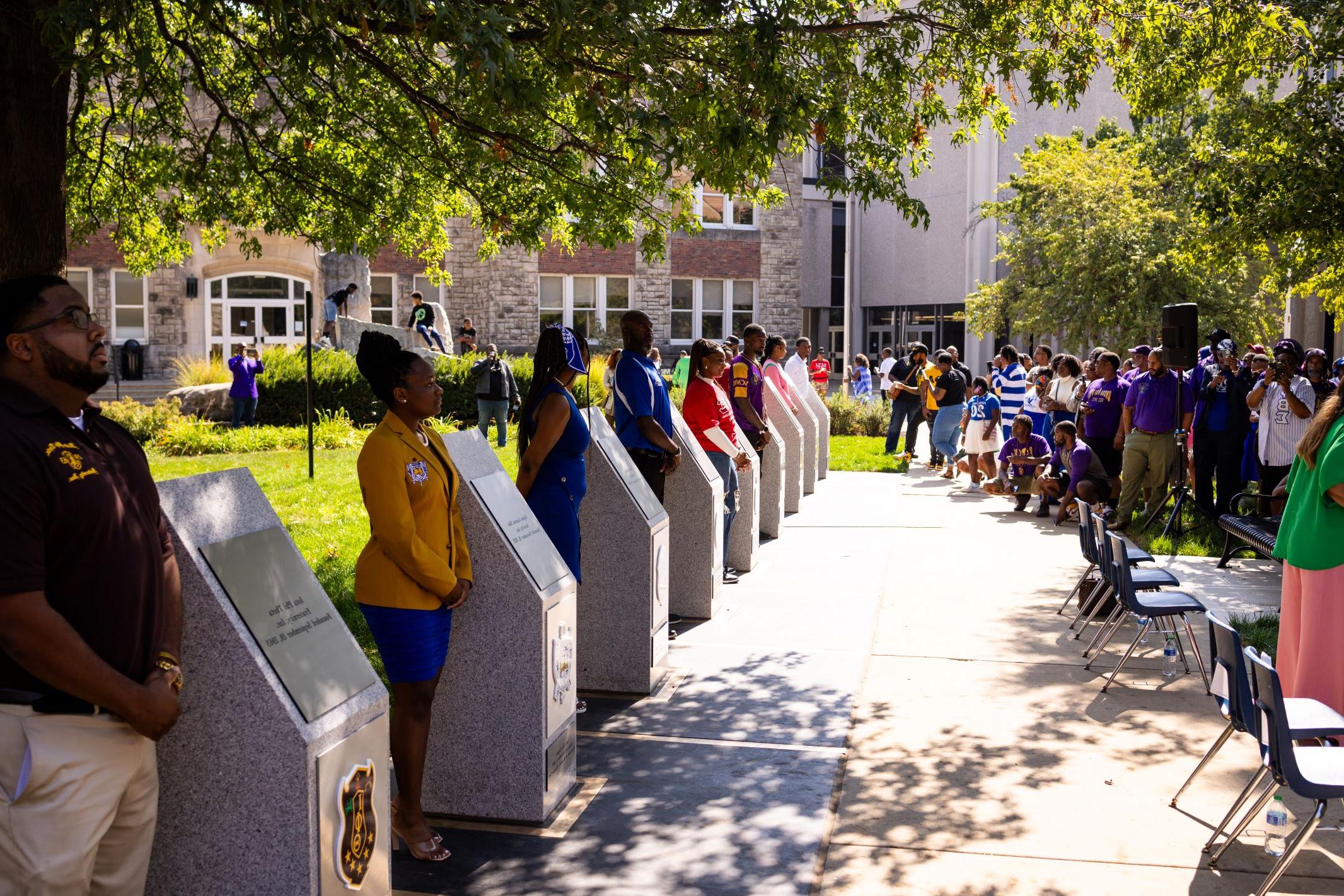 Representatives of the Divine Nine Black Greek organizations stand next to there fraternity's or sorority's plaque.  