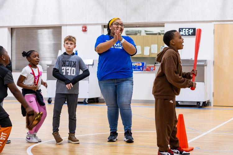 Kayla stands smiling behind students who are lined up to bat inside a gymnasium. The student in front is holding a bright red baseball bat.