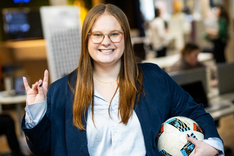 Annmarie Orlando holds a soccer ball under one arm and a roo up gesture with the other hand inside the Sporting KC office